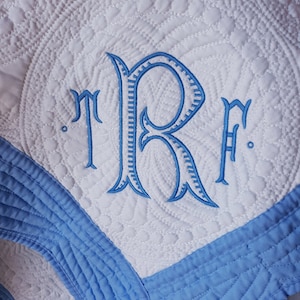 May include: A white quilted blanket featuring a blue embroidered monogram with the letters TRF in a decorative script font. The blanket has a wide blue border and intricate circular stitching patterns throughout the white fabric surface.