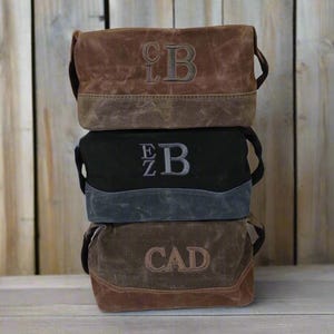 May include: Three stacked brown, black, and gray canvas bags with personalized initials embroidered on the front. Each bag has a contrasting bottom panel and a top handle. The bags are displayed against a wooden background.