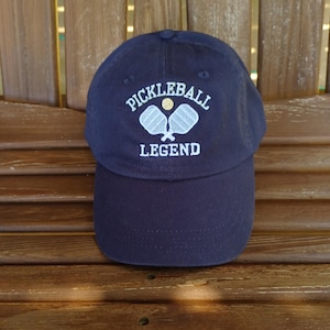 May include: A navy blue baseball cap with white embroidery that reads "Pickleball Legend" and features two white pickleball paddles and a yellow ball.