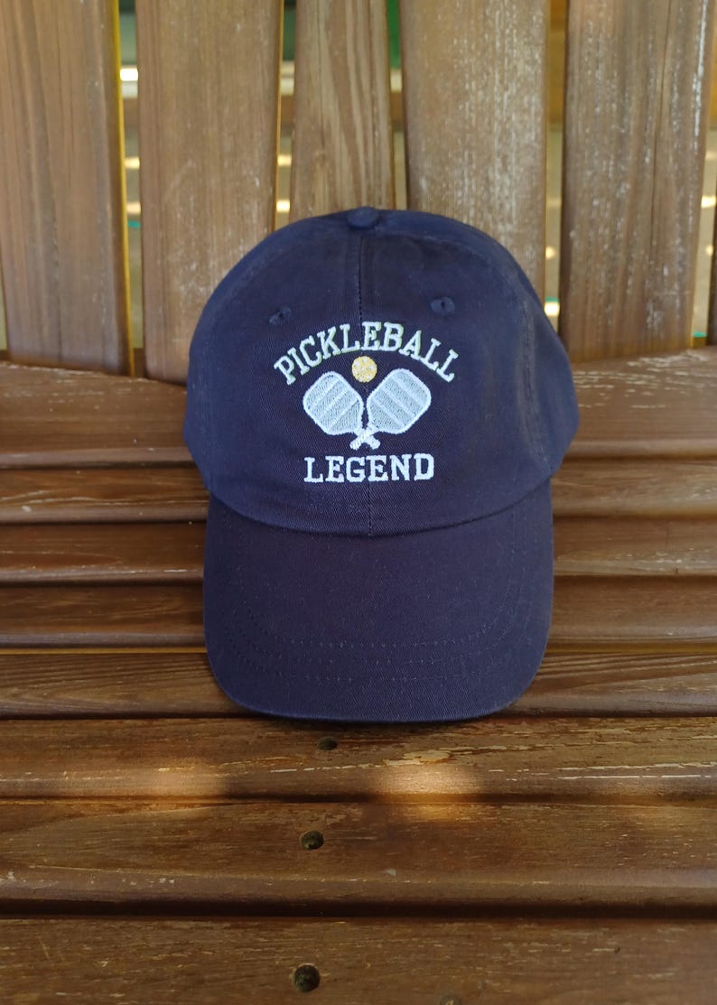 May include: A navy blue baseball cap with white embroidery that reads "Pickleball Legend" and features two white pickleball paddles and a yellow ball.