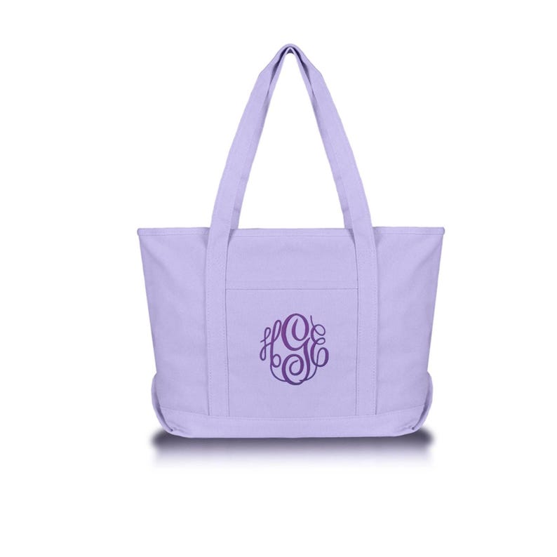 May include: A light lavender canvas tote bag featuring two long shoulder straps and a flat base. The front of the bag displays a dark purple embroidered monogram with the letters HGE in a decorative, swirling script font centered on the pocket.