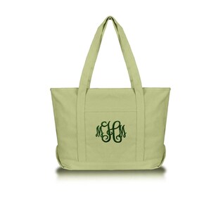 May include: A light sage green canvas tote bag featuring two sturdy shoulder straps and a front pocket. The center of the bag is embroidered with a dark green monogram consisting of the letters M, H, and M in a decorative, swirling script font.