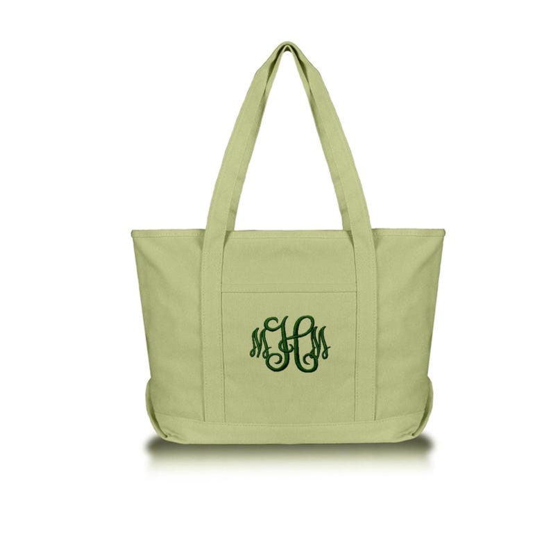 May include: A light sage green canvas tote bag featuring two sturdy shoulder straps and a front pocket. The center of the bag is embroidered with a dark green monogram consisting of the letters M, H, and M in a decorative, swirling script font.