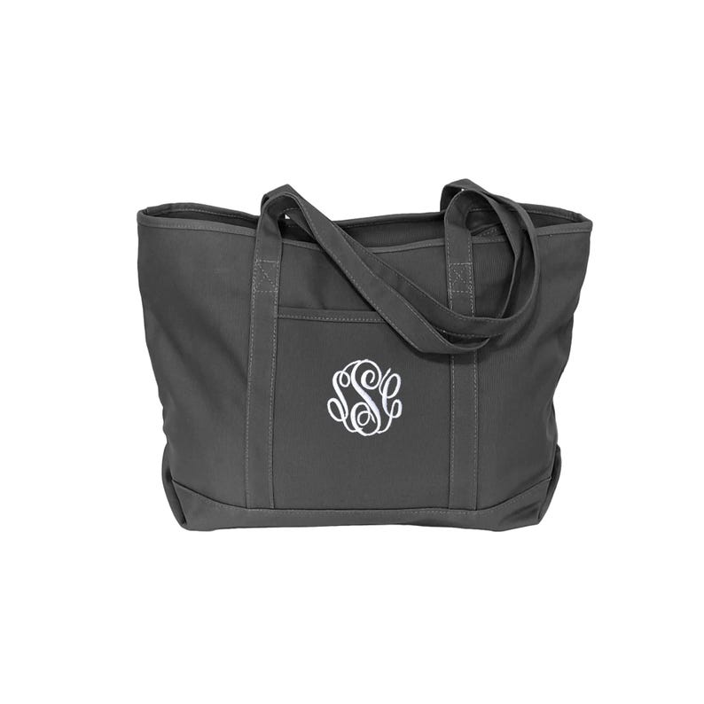 May include: A dark gray canvas tote bag featuring two sturdy shoulder straps and a reinforced bottom panel. The front of the bag displays a white embroidered circular monogram with the letters S, S, and C intertwined in a decorative script font.