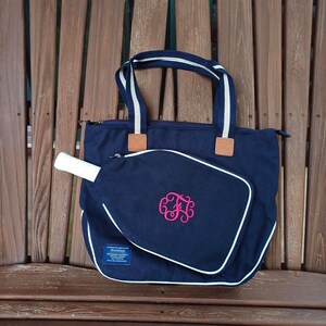 May include: Navy blue tote bag with white and navy striped handles and a matching paddle cover. The paddle cover has a white border and a pink monogram. The bag is sitting on a wooden bench.