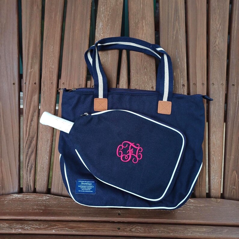 May include: Navy blue tote bag with white and navy striped handles and a matching paddle cover. The paddle cover has a white border and a pink monogram. The bag is sitting on a wooden bench.