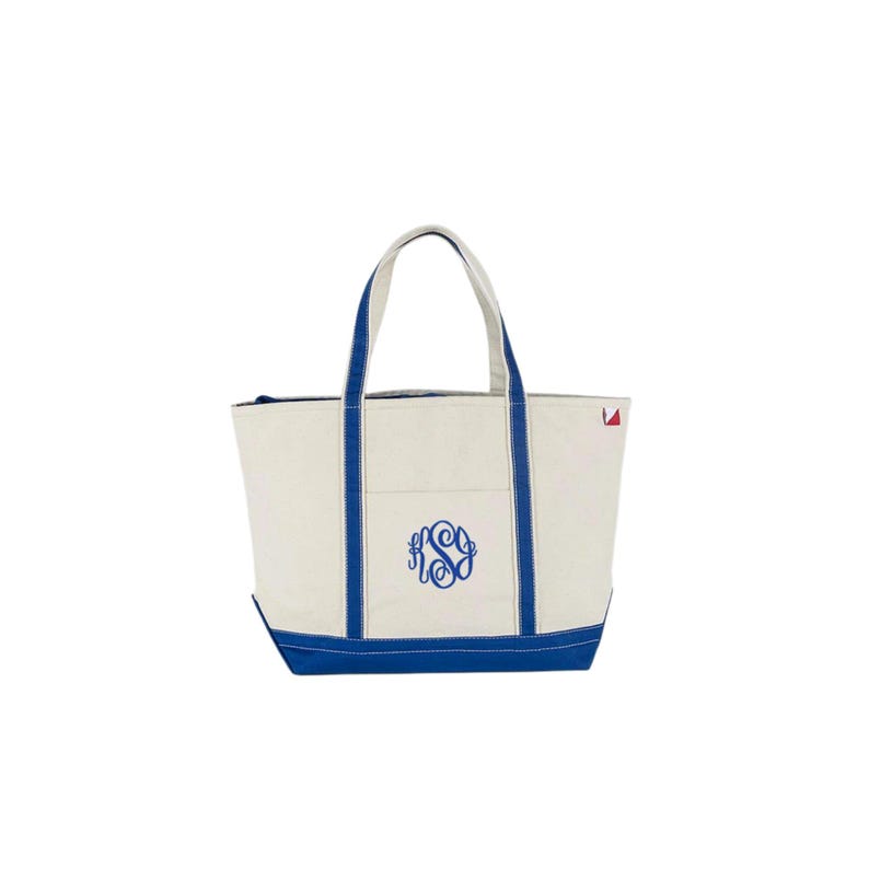 May include: Cream-colored tote bag with blue accents. The bag features blue handles, a blue bottom, and blue trim along the sides. A blue monogram is embroidered on the front. A small red triangle is on the upper right corner.
