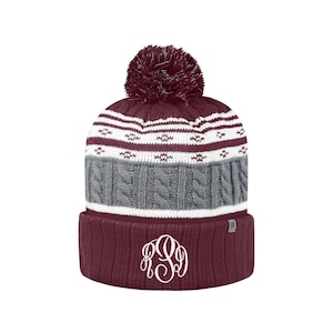 May include: A maroon knit beanie with a white pom pom on top. The beanie has a white and gray cable knit pattern and a white band around the bottom. The beanie has a white monogram on the front.