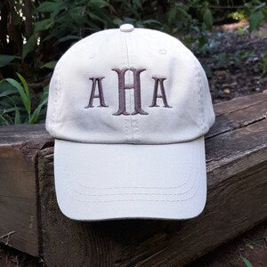 May include: A light beige baseball cap with the embroidered letters "AHA" in a darker brown color. The cap is sitting on a weathered wooden surface, with greenery in the background.