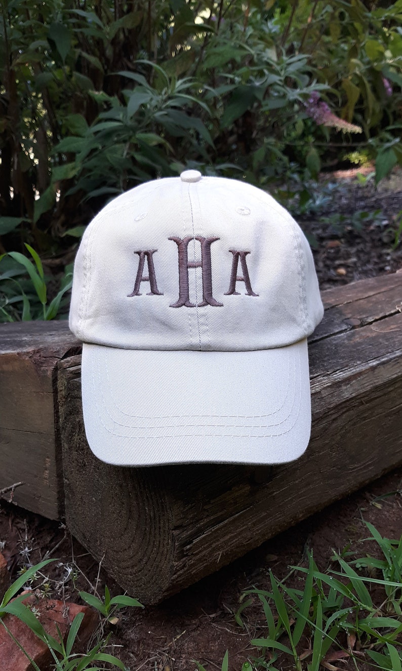 May include: A light beige baseball cap with the embroidered letters "AHA" in a darker brown color. The cap is sitting on a weathered wooden surface, with greenery in the background.