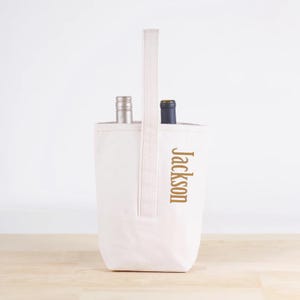 May include: A white canvas wine tote with a handle, holding two wine bottles. The word "Jackson" is printed vertically in gold on the front of the tote. The tote is sitting on a wooden surface.