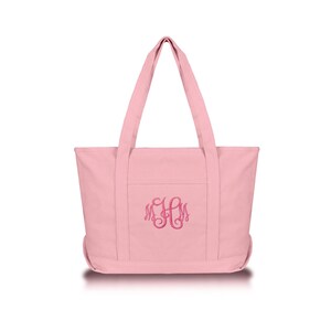 May include: A light pink canvas tote bag featuring two sturdy shoulder straps and a front pocket. The center of the bag is embroidered with a dark pink monogram featuring the letters M, H, and M in a decorative, swirling cursive script font.