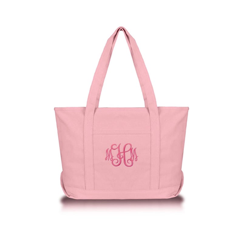 May include: A light pink canvas tote bag featuring two sturdy shoulder straps and a front pocket. The center of the bag is embroidered with a dark pink monogram featuring the letters M, H, and M in a decorative, swirling cursive script font.