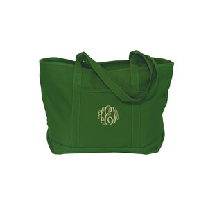 May include: A forest green canvas tote bag featuring two sturdy shoulder straps and an exterior slip pocket. The center of the bag is embroidered with a decorative monogram featuring the letters E, H, and M in a light beige thread color.
