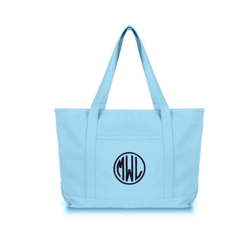 May include: A light blue canvas tote bag featuring two sturdy shoulder straps and a front exterior pocket. The center of the bag displays a circular navy blue embroidered monogram with the letters MWL enclosed within the circular border.