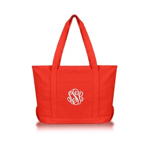 May include: A bright red canvas tote bag featuring two long shoulder straps and a centered white embroidered monogram with the letters S, S, and C in a decorative script font. The bag has a rectangular body with a flat base and vertical stitching.