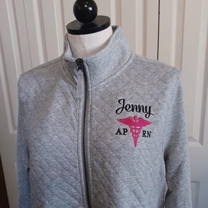 May include: A light gray quilted zip-up jacket featuring a high collar and a black zipper. Embroidered on the left chest is the name Jenny in black script, above a pink caduceus symbol with the black letters AP and RN on either side.