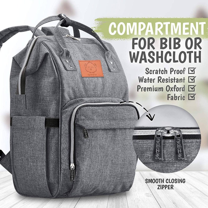 K&ouml;nnte beinhalten: Grauer Wickeltasche-Rucksack mit einem Rei&szlig;verschlussfach mit der Aufschrift "COMPARTMENT FOR BIB OR WASHCLOTH". Die Tasche hat ein Rei&szlig;verschlussfach vorne und ein gro&szlig;es Fach an der Seite. Die Tasche ist aus wasserabweisendem, kratzfestem Stoff.