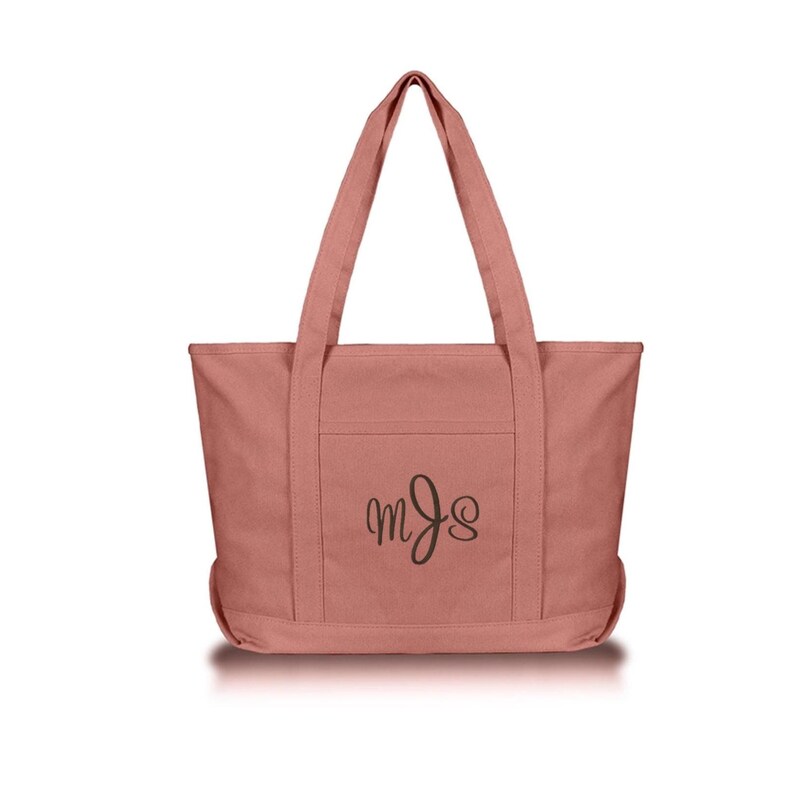 May include: A dusty rose canvas tote bag featuring two sturdy shoulder straps and a front exterior pocket. The center of the front pocket displays a dark brown embroidered monogram with the stylized cursive letters M, J, and S in a vertical arrangement.