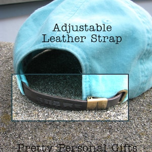 May include: A light blue baseball cap with an adjustable brown leather strap and a brass buckle. The text "Adjustable Leather Strap" is visible on the cap. The words "Pretty Personal Gifts" are also visible.