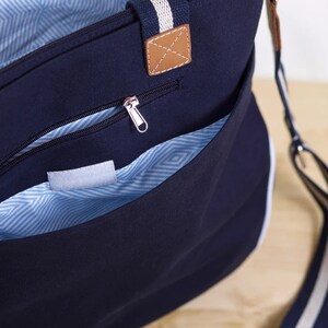 May include: Navy blue tote bag with white trim and a striped strap. The bag features a zippered pocket and a front pocket. The interior lining has a blue and white geometric pattern. A brown leather tag is attached to the handle.