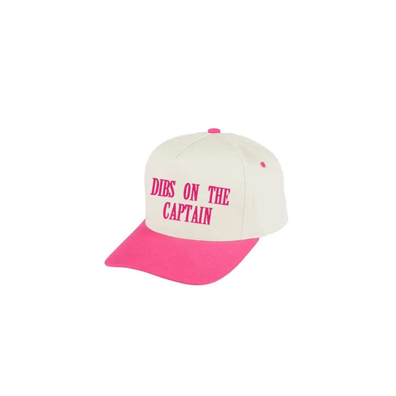 K&ouml;nnte beinhalten: Baseballkappe mit wei&szlig;er Krone und pinkfarbenem Schirm. Der Text "DIBS ON THE CAPTAIN" ist in Pink auf die Vorderseite der Kappe gedruckt. Die Kappe hat einen pinkfarbenen Knopf oben.