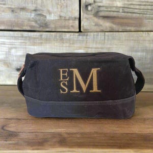 May include: A dark charcoal gray rectangular toiletry bag featuring a structured design with a reinforced base. The front of the bag is embroidered with the large gold-colored letters E and M, with the letter S positioned above the letter E.
