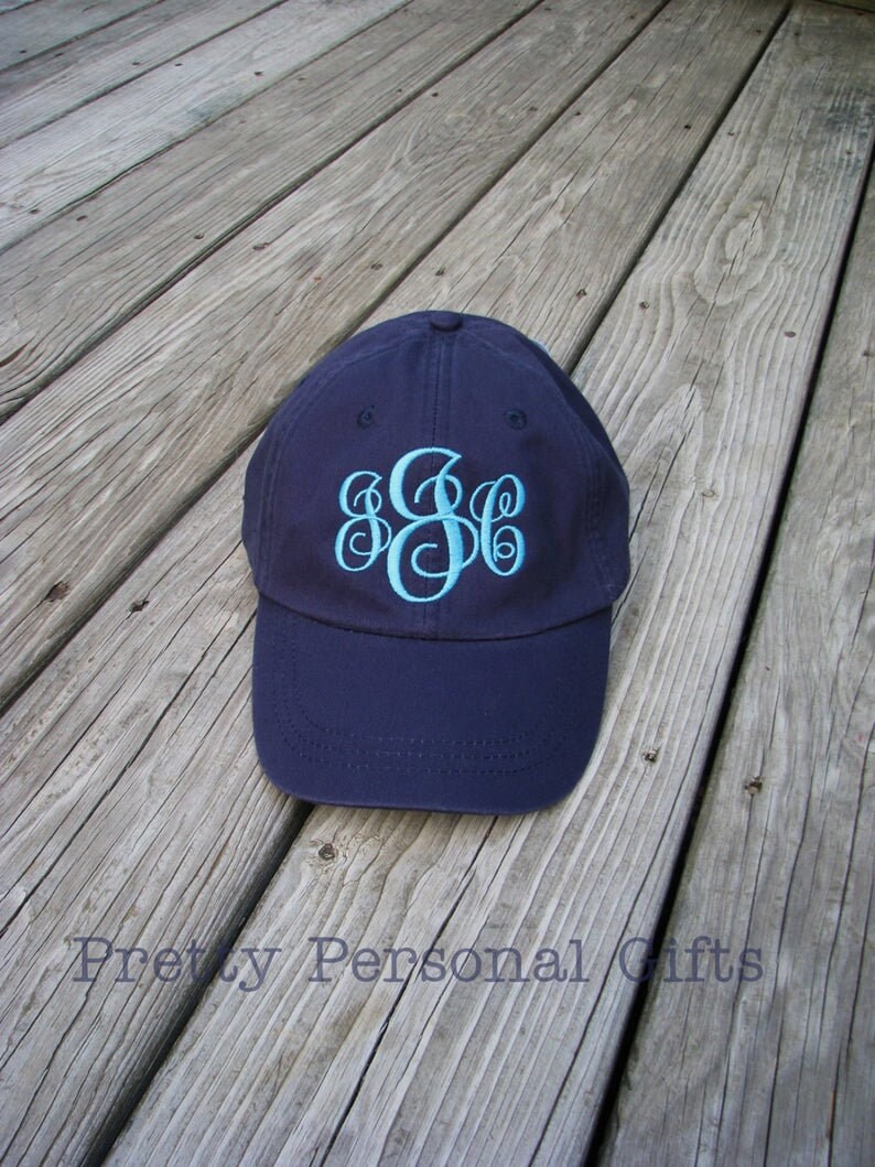 May include: Navy blue baseball cap with a light blue monogram. The cap is sitting on a weathered wooden surface. The text "Pretty Personal Gifts" is visible at the bottom of the image.