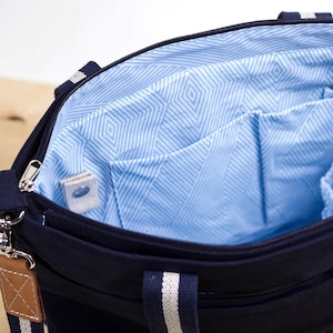 May include: Navy blue tote bag with a light blue geometric patterned interior. The bag has a zipper closure, multiple interior pockets, and a detachable shoulder strap. The handles and strap feature white stripes.