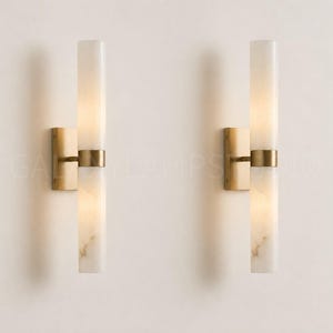 Puede incluir: Un par de apliques de pared con pantallas cilíndricas de alabastro blanco con vetas naturales, sujetas por una banda central de latón cepillado y una placa de montaje rectangular de latón. El diseño incluye dos segmentos de piedra apilados.
