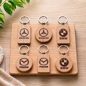 Könnte beinhalten: Sechs Holzschlüsselanhänger mit Metallringen, angeordnet in zwei Reihen. Die Schlüsselanhänger zeigen lasergravierte Logos von Mercedes-Benz, BMW und Mazda in rechteckigen und runden Formen, die eine natürliche Holzmaserung aufweisen.