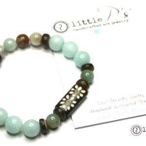 May include: A beaded bracelet featuring a mix of light blue, brown, and white beads. A dark brown cylindrical bead with a white floral design is a focal point. The bracelet is on a white background.