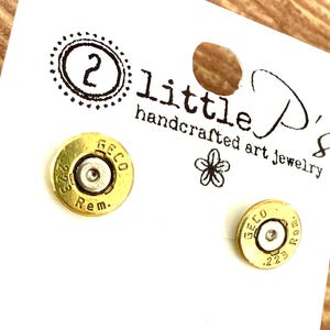 May include: A pair of handcrafted art jewelry earrings made from .223 Rem brass casings. The earrings are gold-colored with silver-colored centers, and are displayed on a white card with the text "little P's handcrafted art jewelry".