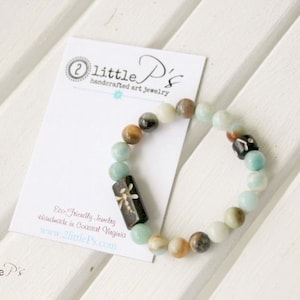 May include: A light blue and brown beaded bracelet with a dragonfly charm. The bracelet is handmade and eco-friendly. The text on the card reads "little P's handcrafted art jewelry".