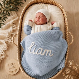 Può includere: Un neonato con berretto e maglione in maglia bianca, che riposa in una cesta intrecciata. Il bambino è coperto da una coperta in maglia azzurra con il nome liam ricamato in corsivo bianco. Un disco di legno con la scritta hello world è vicino.