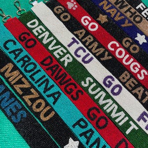 May include: A collection of beaded lanyards featuring various team names and slogans including Go Navy, Go Army, Go Cougs, Denver, TCU, Summit, Go Dawgs, Carolina, Mizzou, and Panthers. Each strap is crafted with colorful glass beads and a metal clasp.
