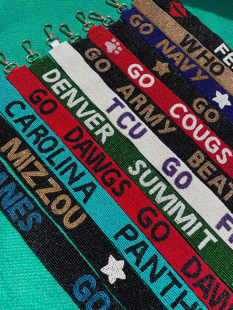 May include: A collection of beaded lanyards featuring various team names and slogans including Go Navy, Go Army, Go Cougs, Denver, TCU, Summit, Go Dawgs, Carolina, Mizzou, and Panthers. Each strap is crafted with colorful glass beads and a metal clasp.