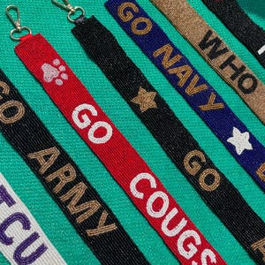 May include: A collection of beaded lanyards featuring various designs and text including GO ARMY, GO COUGS, GO NAVY, and WHO. Each strap is crafted with small glass beads in colors like red, black, blue, white, and gold, finished with a metal lobster clasp.