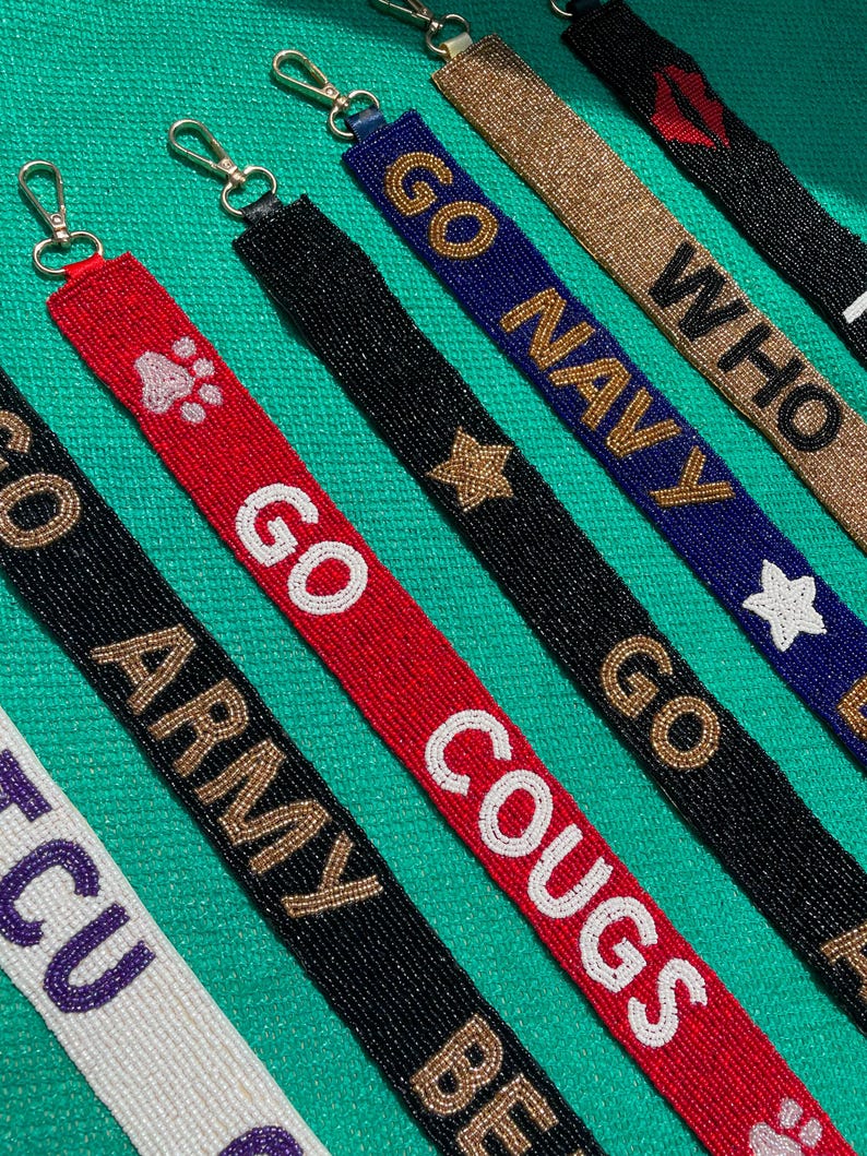 May include: A collection of beaded lanyards featuring various designs and text including GO ARMY, GO COUGS, GO NAVY, and WHO. Each strap is crafted with small glass beads in colors like red, black, blue, white, and gold, finished with a metal lobster clasp.