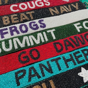 May include: A collection of rectangular beaded wristlet keychains in various colors including red, black, white, green, and teal. Each strap features embroidered text such as BEAT ARMY, COUGS, BEAT NAVY, FROGS, SUMMIT, GO DAWGS, PANTHERS, and MIZZOU.
