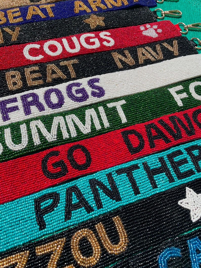 May include: A collection of rectangular beaded wristlet keychains in various colors including red, black, white, green, and teal. Each strap features embroidered text such as BEAT ARMY, COUGS, BEAT NAVY, FROGS, SUMMIT, GO DAWGS, PANTHERS, and MIZZOU.