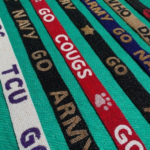 May include: A collection of long, narrow beaded straps featuring various text and symbols. The straps include white with purple text, black with gold text, red with white text, and blue with gold text. Phrases include TCU GO, BEAT NAVY, GO COUGS, ARMY, BEAT ARMY, and JAZZ.