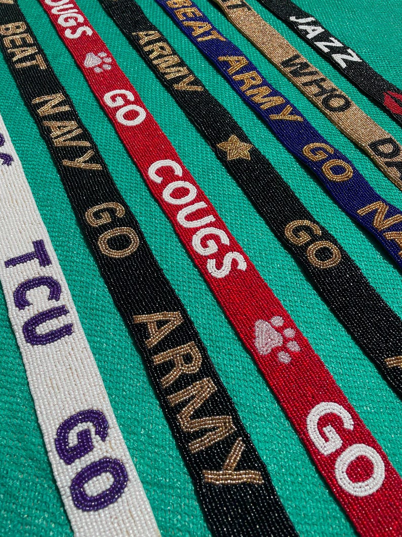 May include: A collection of long, narrow beaded straps featuring various text and symbols. The straps include white with purple text, black with gold text, red with white text, and blue with gold text. Phrases include TCU GO, BEAT NAVY, GO COUGS, ARMY, BEAT ARMY, and JAZZ.