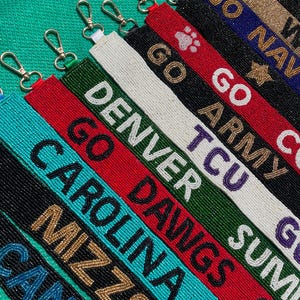 May include: A collection of rectangular beaded keychains featuring various team names and logos including Denver, Carolina, Mizzou, TCU, Go Army, and Go Navy. Each strap is crafted with colorful seed beads and includes a silver-toned metal lobster clasp.