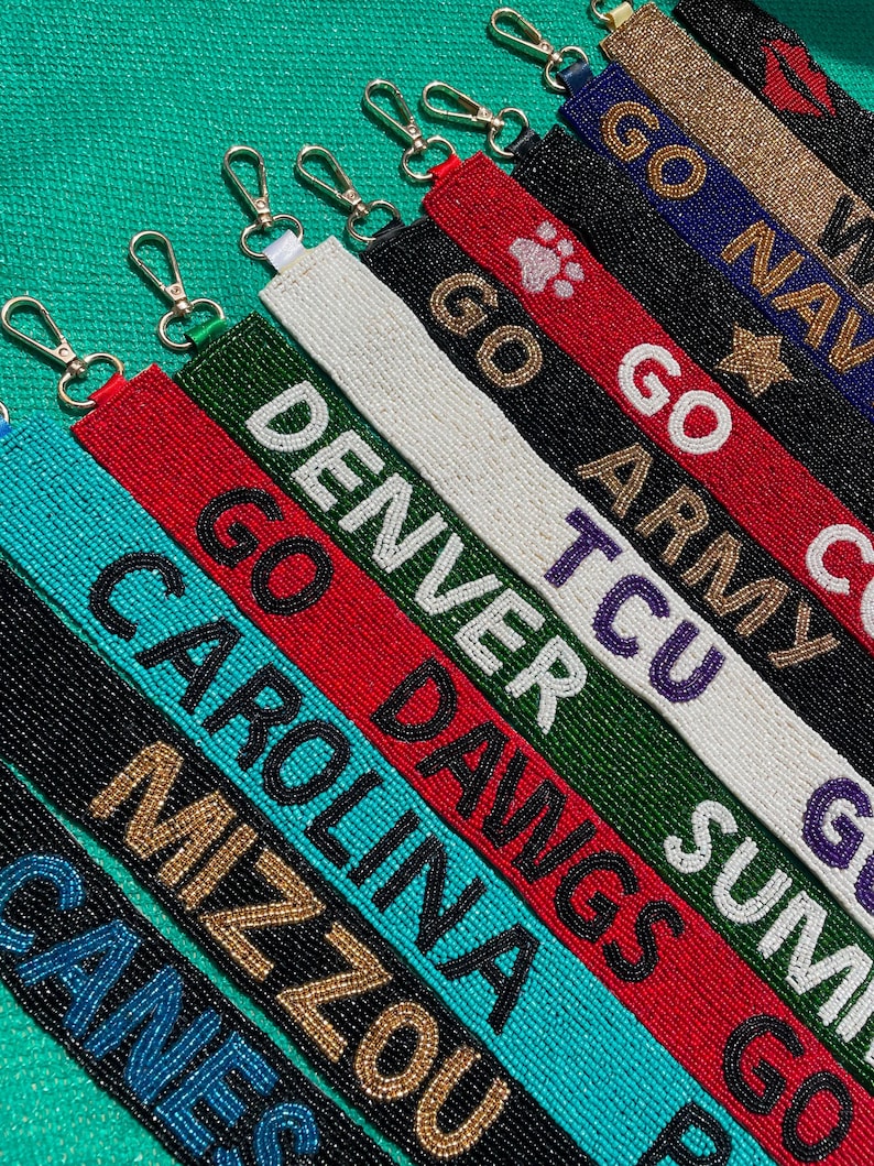 May include: A collection of rectangular beaded keychains featuring various team names and logos including Denver, Carolina, Mizzou, TCU, Go Army, and Go Navy. Each strap is crafted with colorful seed beads and includes a silver-toned metal lobster clasp.
