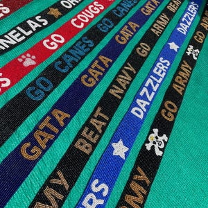 May include: A collection of narrow, beaded straps in various colors including red, black, blue, and navy. Each strap features embroidered text and symbols such as GIG, ORNELAS, GO COUGS, GO CANES, GATA, BEAT NAVY, DAZZLERS, and GO ARMY with star and paw icons.