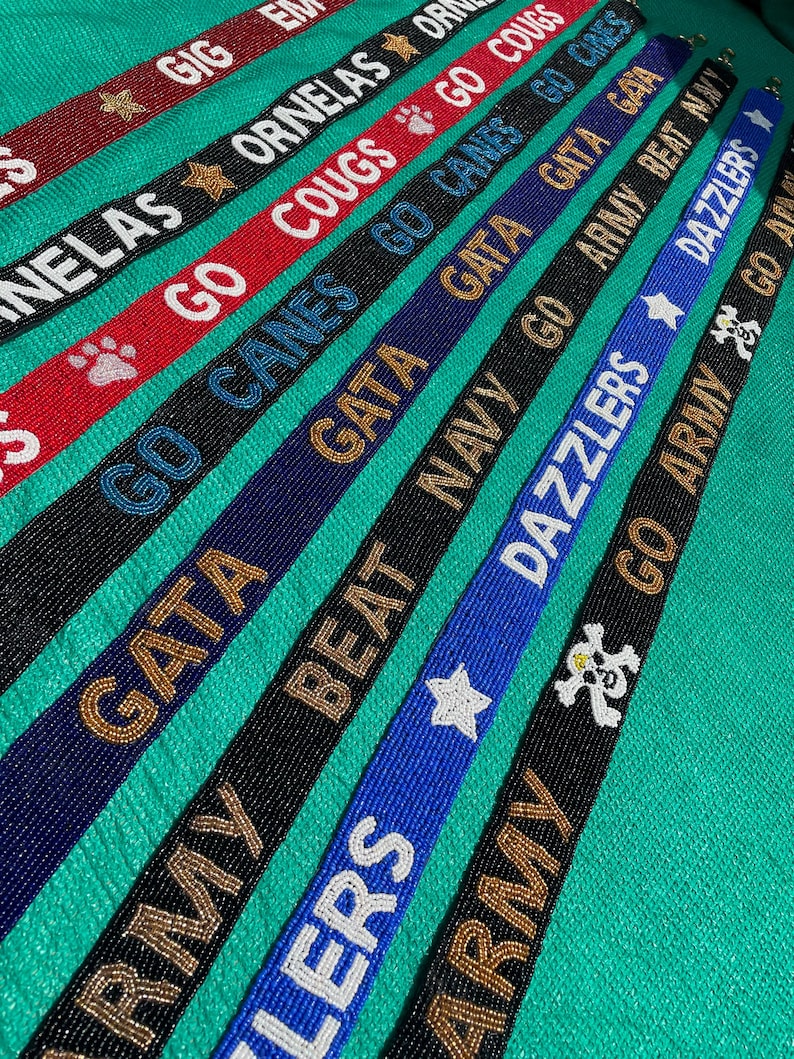 May include: A collection of narrow, beaded straps in various colors including red, black, blue, and navy. Each strap features embroidered text and symbols such as GIG, ORNELAS, GO COUGS, GO CANES, GATA, BEAT NAVY, DAZZLERS, and GO ARMY with star and paw icons.