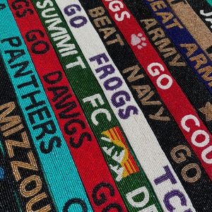 May include: A collection of narrow, rectangular beaded straps arranged in parallel rows. Each strap features distinct color combinations and text including MIZZOU, PANTHERS, GO DAWGS, SUMMIT FC, GO FROGS, GO NAVY, BEAT ARMY, and GIMME DAT, with various team-themed icons.