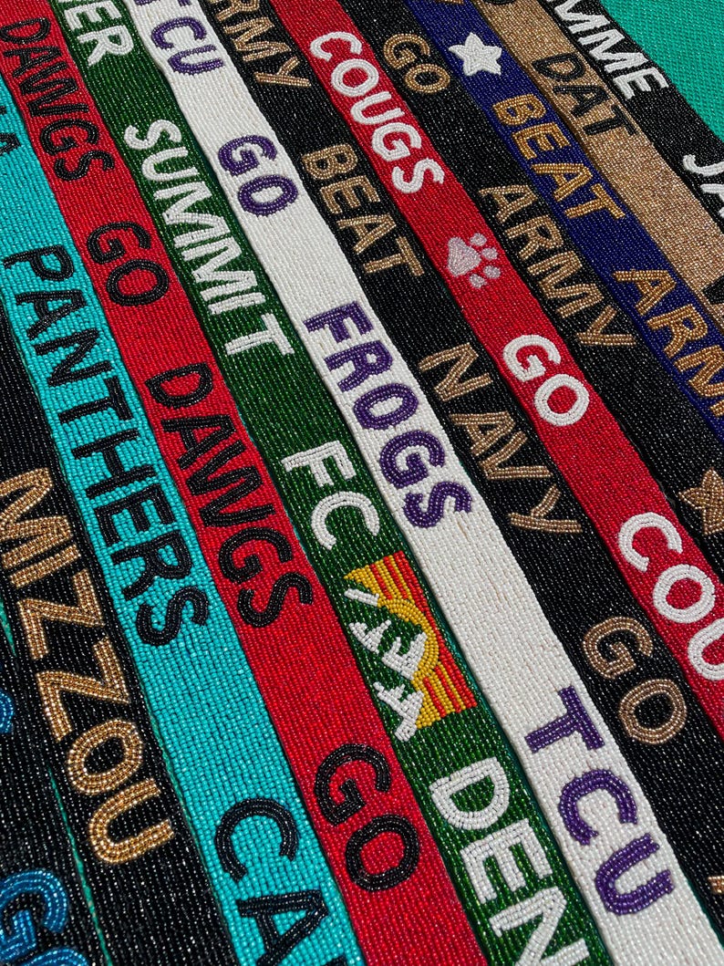 May include: A collection of narrow, rectangular beaded straps arranged in parallel rows. Each strap features distinct color combinations and text including MIZZOU, PANTHERS, GO DAWGS, SUMMIT FC, GO FROGS, GO NAVY, BEAT ARMY, and GIMME DAT, with various team-themed icons.