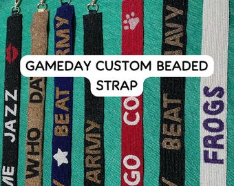 May include: A collection of seven long, narrow beaded straps featuring various colors and text designs including JAZZ, WHO DAT, BEAT ARMY, GO CO, BEAT NAVY, and FROGS. Each strap is finished with a metal lobster clasp at the top for attachment.